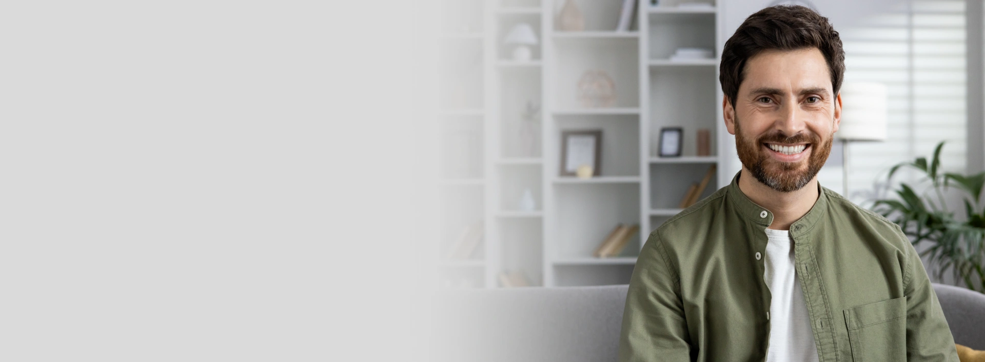Smiling male dentist in olive shirt standing in modern dental office with shelving and plant behind him.